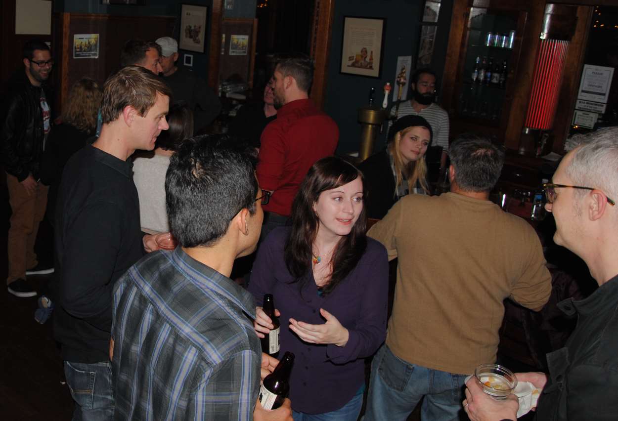 People socializing in a dimly lit bar. Several people holding drinks, engaged in conversation.