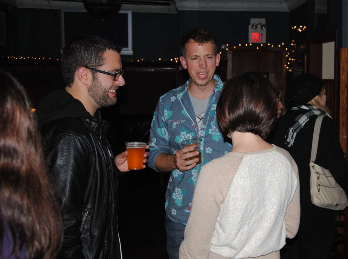 Three people in a bar, two men holding drinks, talking with a woman in a cream sweater. Low lighting.