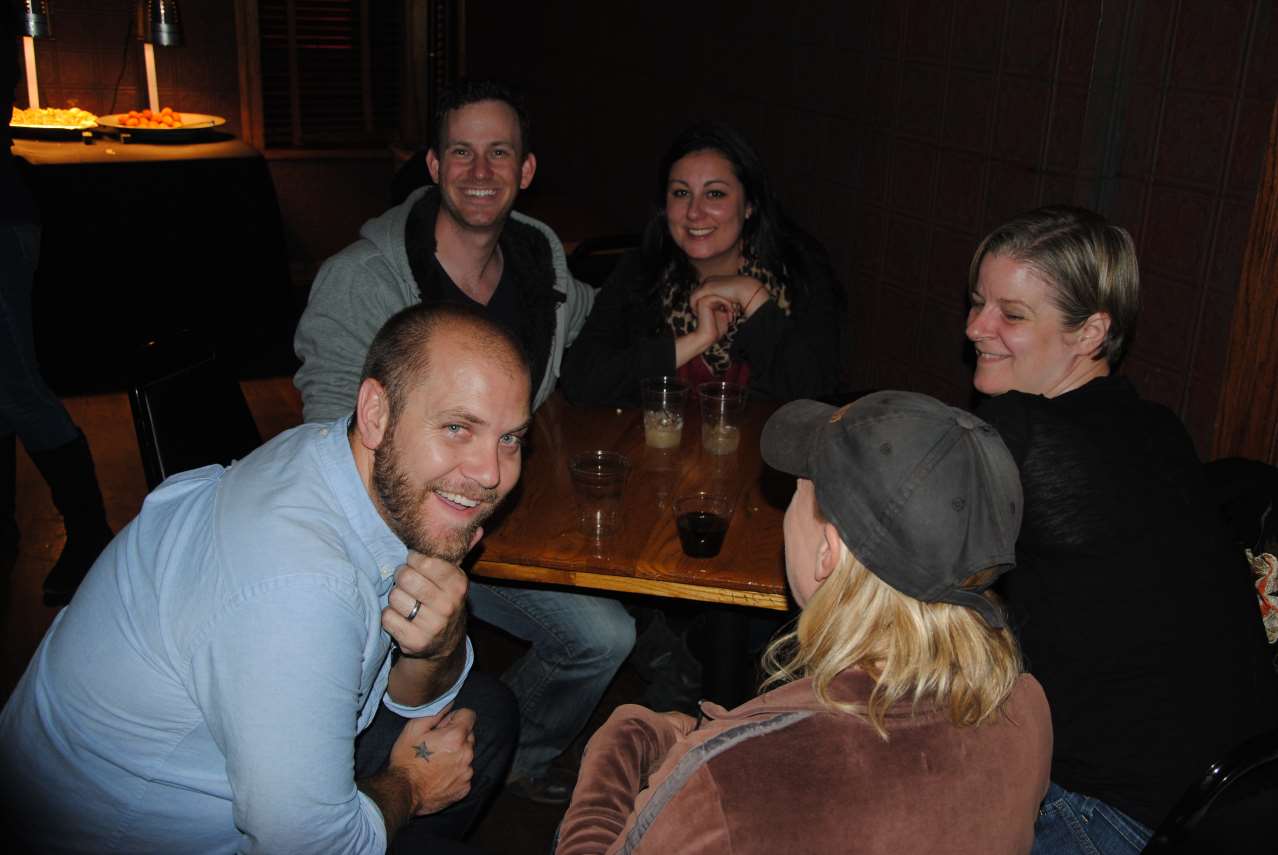 Five people at a wooden table in a dimly lit room, smiling and laughing. One man looks toward the camera.