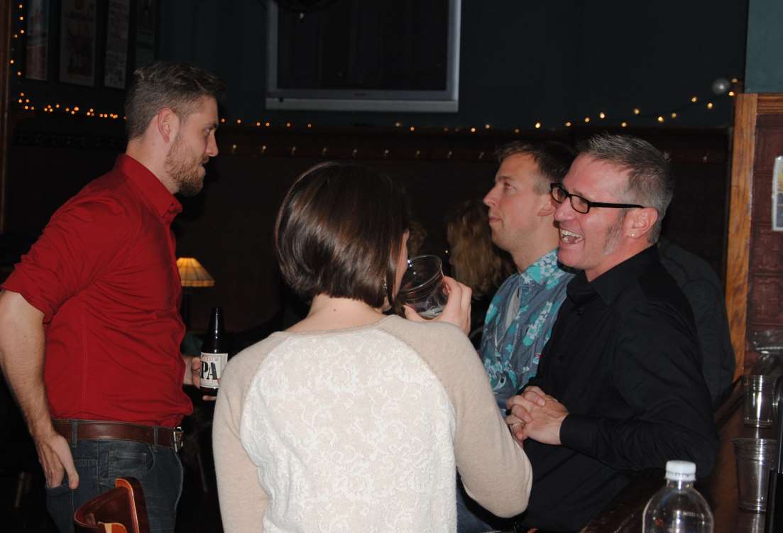People socializing at a bar. Man in red shirt talking to others; man in black shirt smiles.