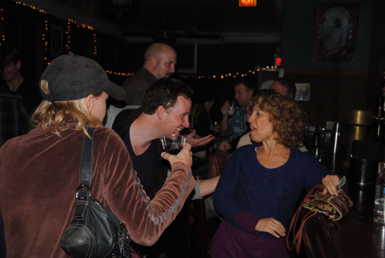 People conversing in a dimly lit bar. Woman in brown velvet coat and hat, man holding a glass.