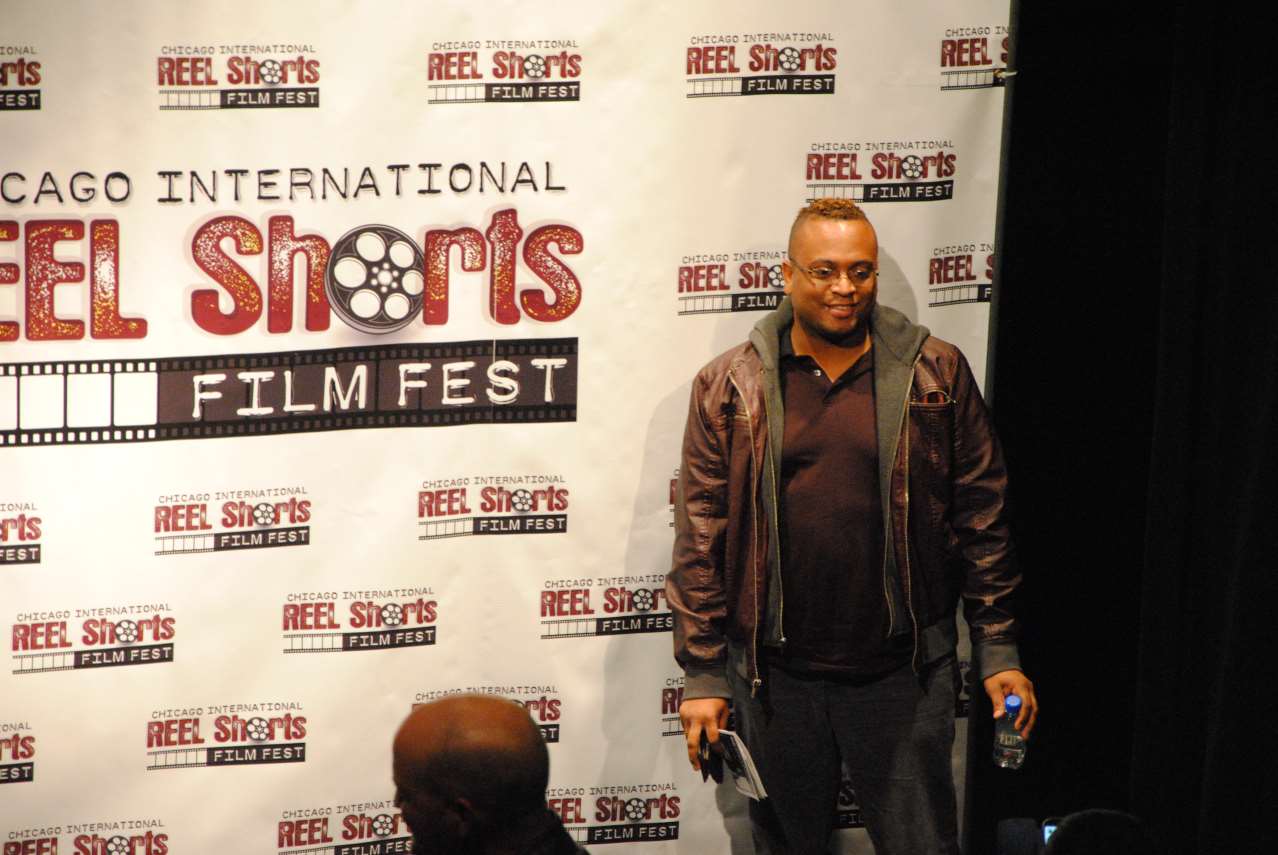 Man at Chicago International Reel Shorts Film Fest. He's wearing a brown jacket, and standing near a backdrop.