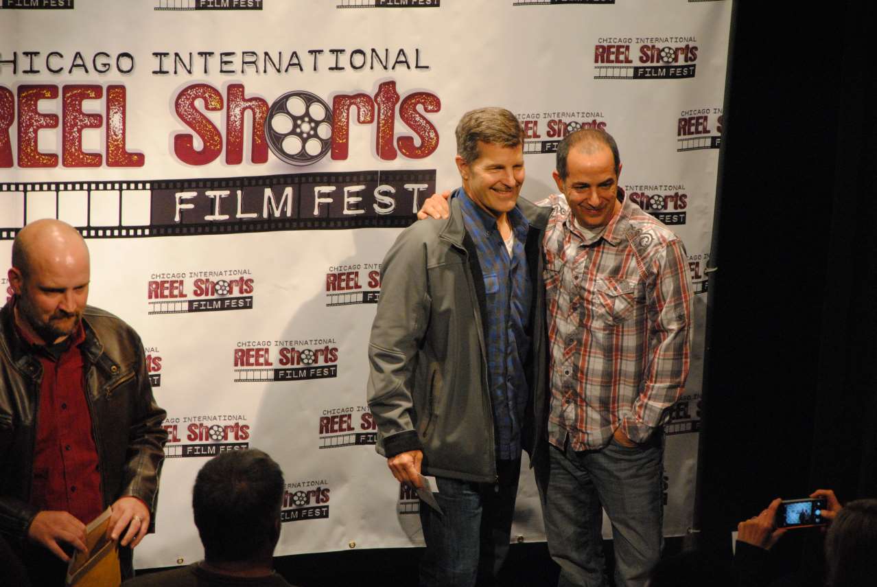 Three men at the Chicago International Reel Shorts Film Fest. Two have arms around each other, smiling in front of a banner.