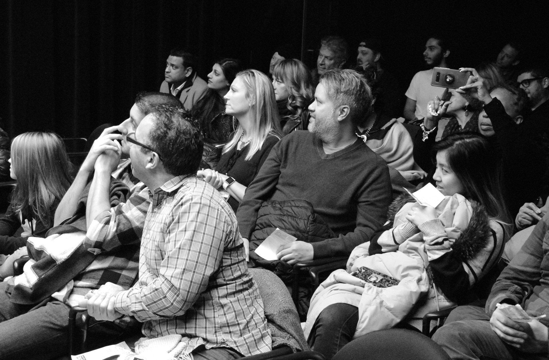 Audience seated in dark theater, faces turned toward unseen stage. Mostly adults, some with programs.