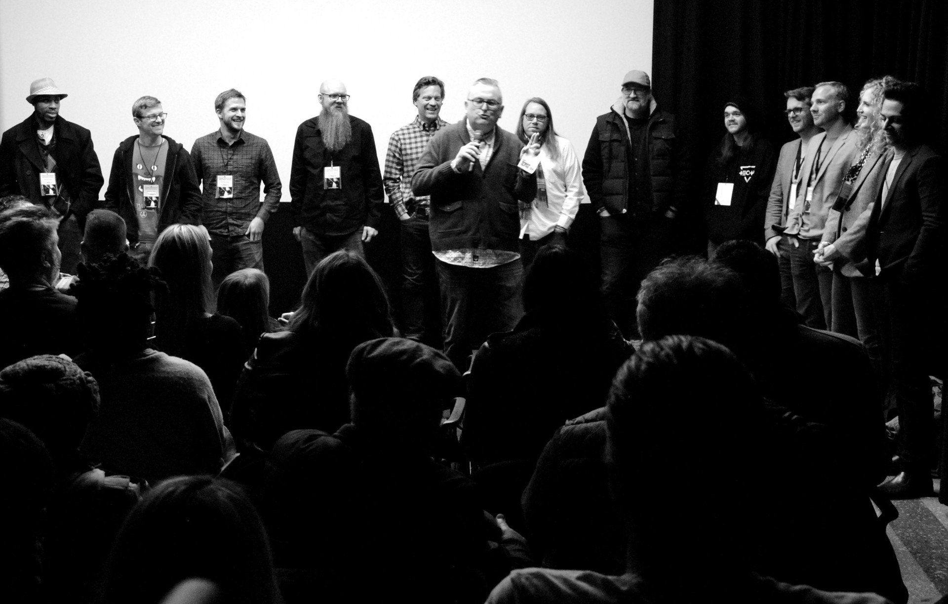 Group of people on stage, facing an audience in a dark theater. One man speaks into a microphone. Black and white.