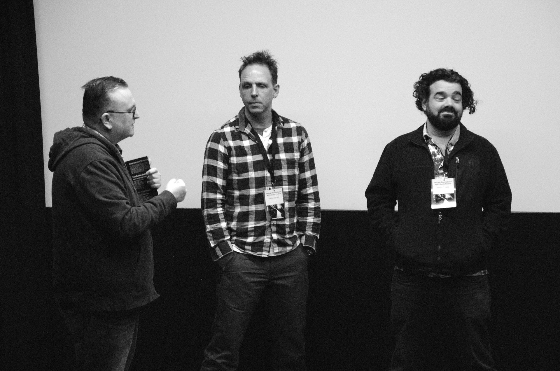 Three men on a stage. A man on the left holds a microphone; two men stand between him and a screen.