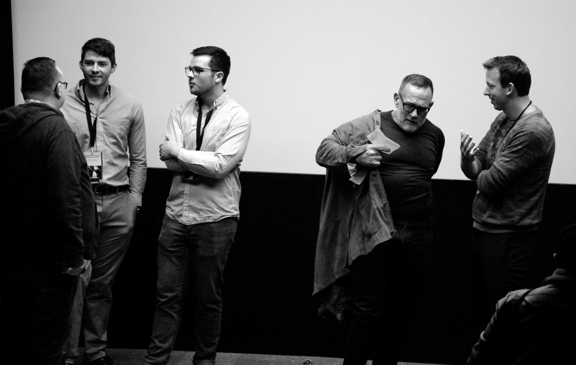 Five men stand in front of a screen. One man takes off a jacket while others talk. Black and white.
