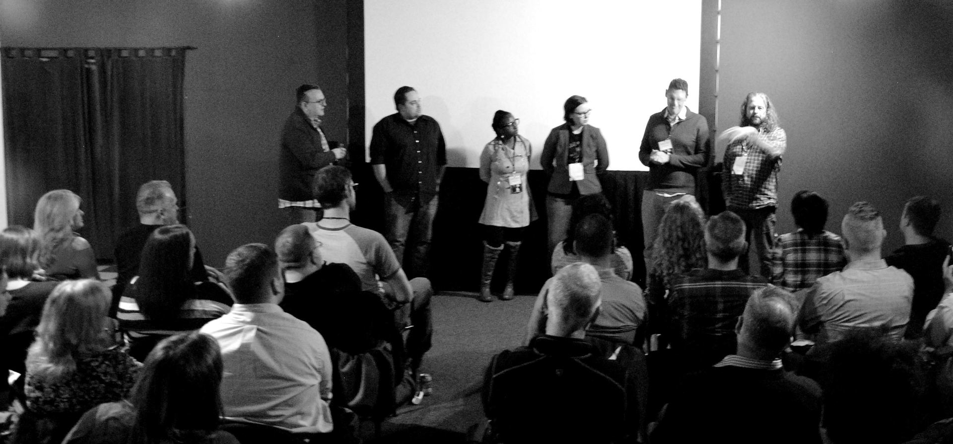 Panel of six people on a stage in front of an audience. Dark room, monochrome photo.