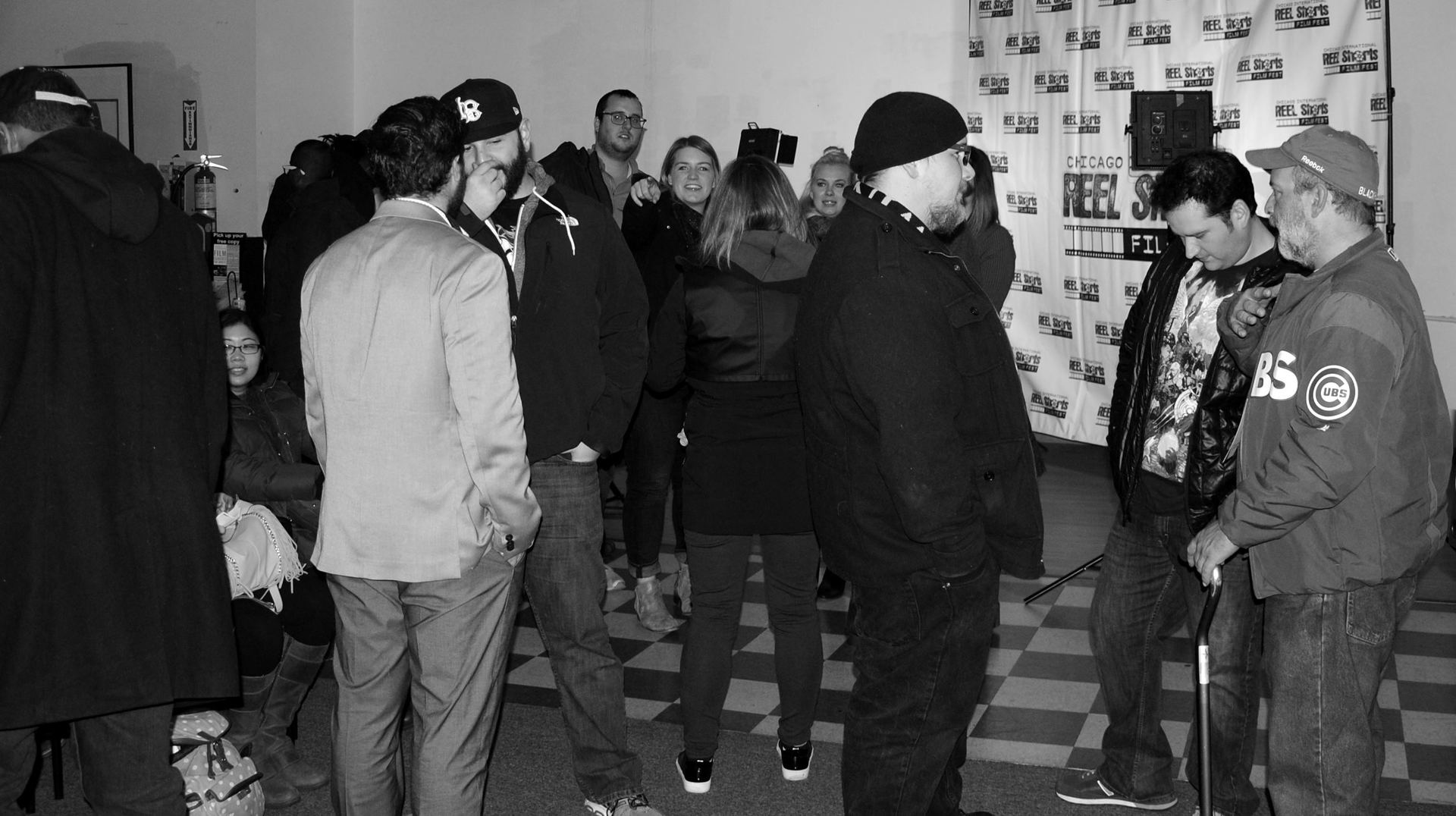 People in a room, some wearing jackets and hats. They stand and talk near a backdrop.