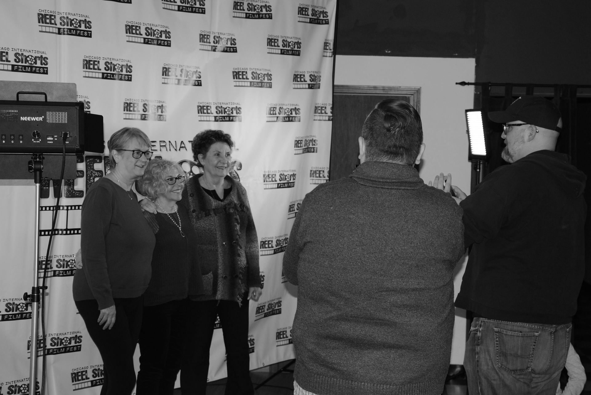 Four women pose for a photo, two men stand with cameras, in front of a banner.