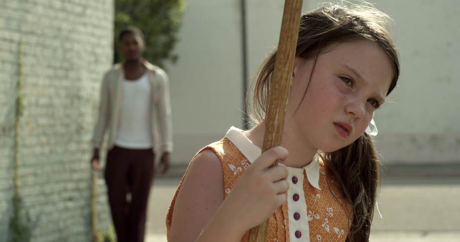 Girl in orange dress holds a stick, looking concerned. Man walks in background, near a brick wall.