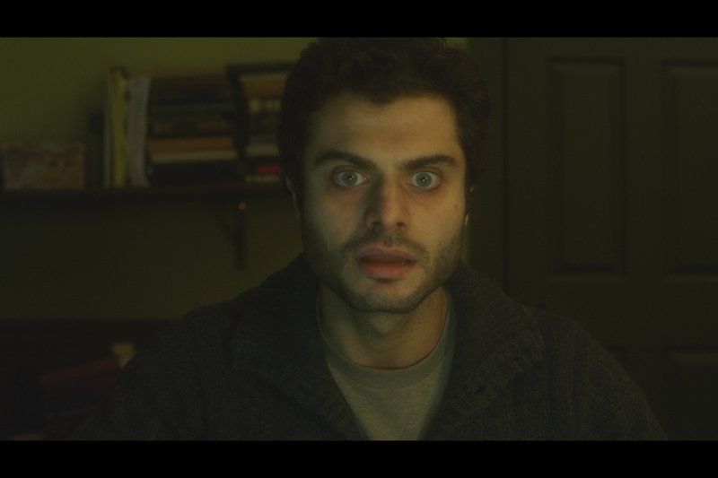 Man with wide eyes and shocked expression stares forward in dimly lit room.