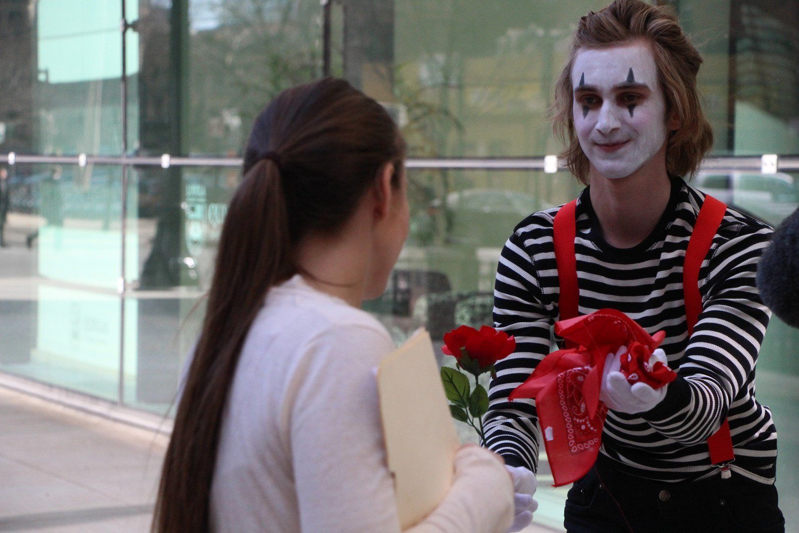 A mime offers a red rose to a woman with long brown hair, indoors.