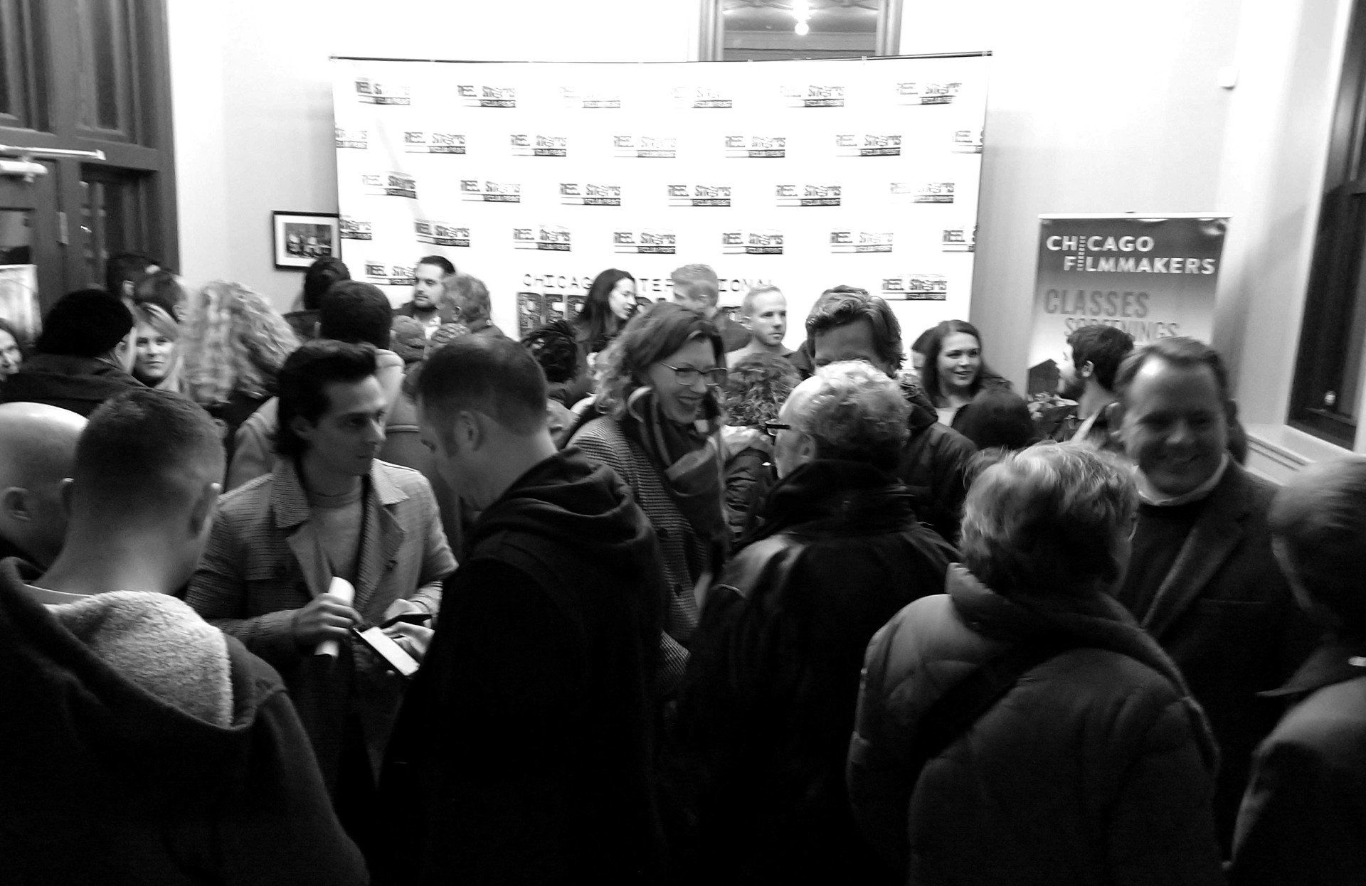 People gather at event with step-and-repeat backdrop and poster; black and white photo.
