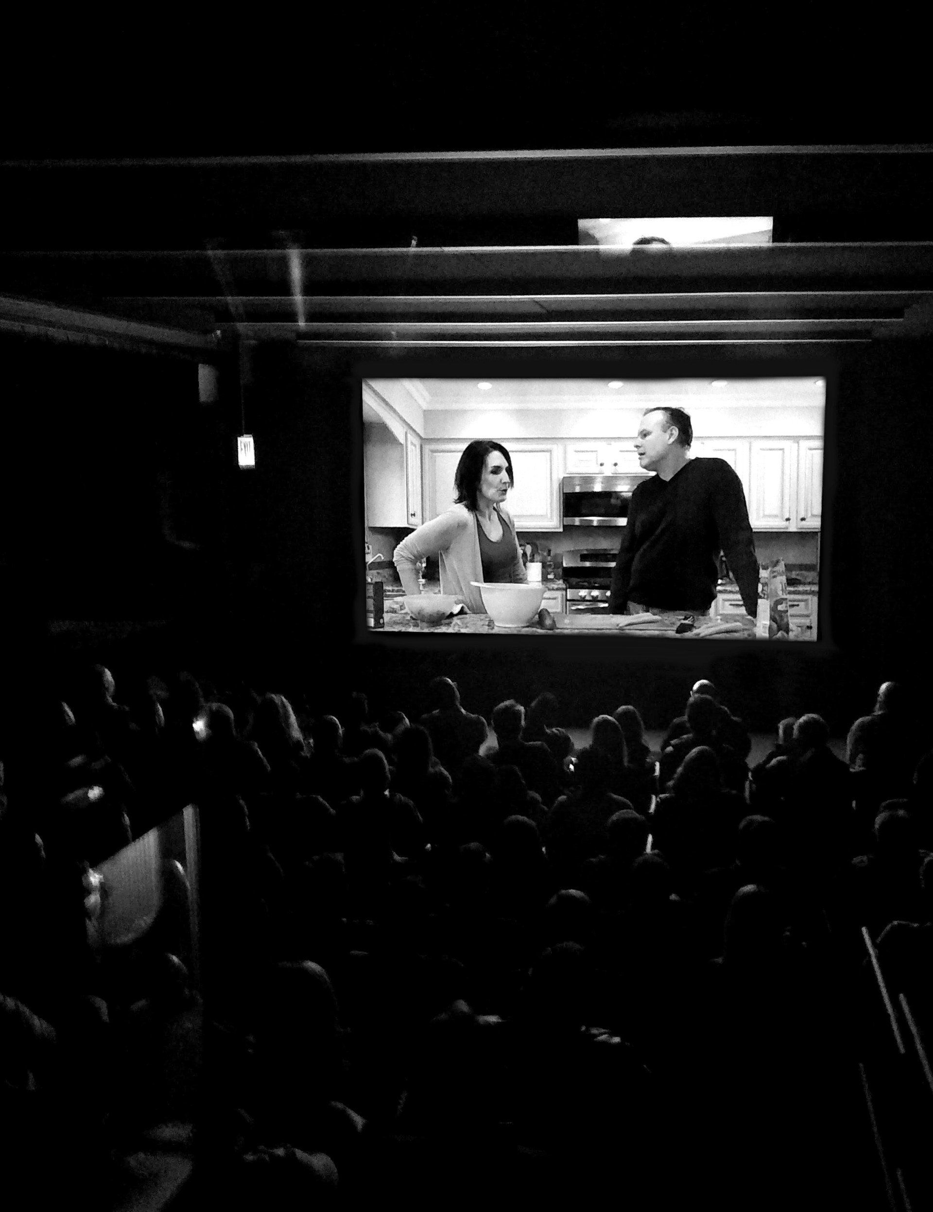 Audience watches a black and white movie of a couple in a kitchen on a large screen in a dimly lit theater.