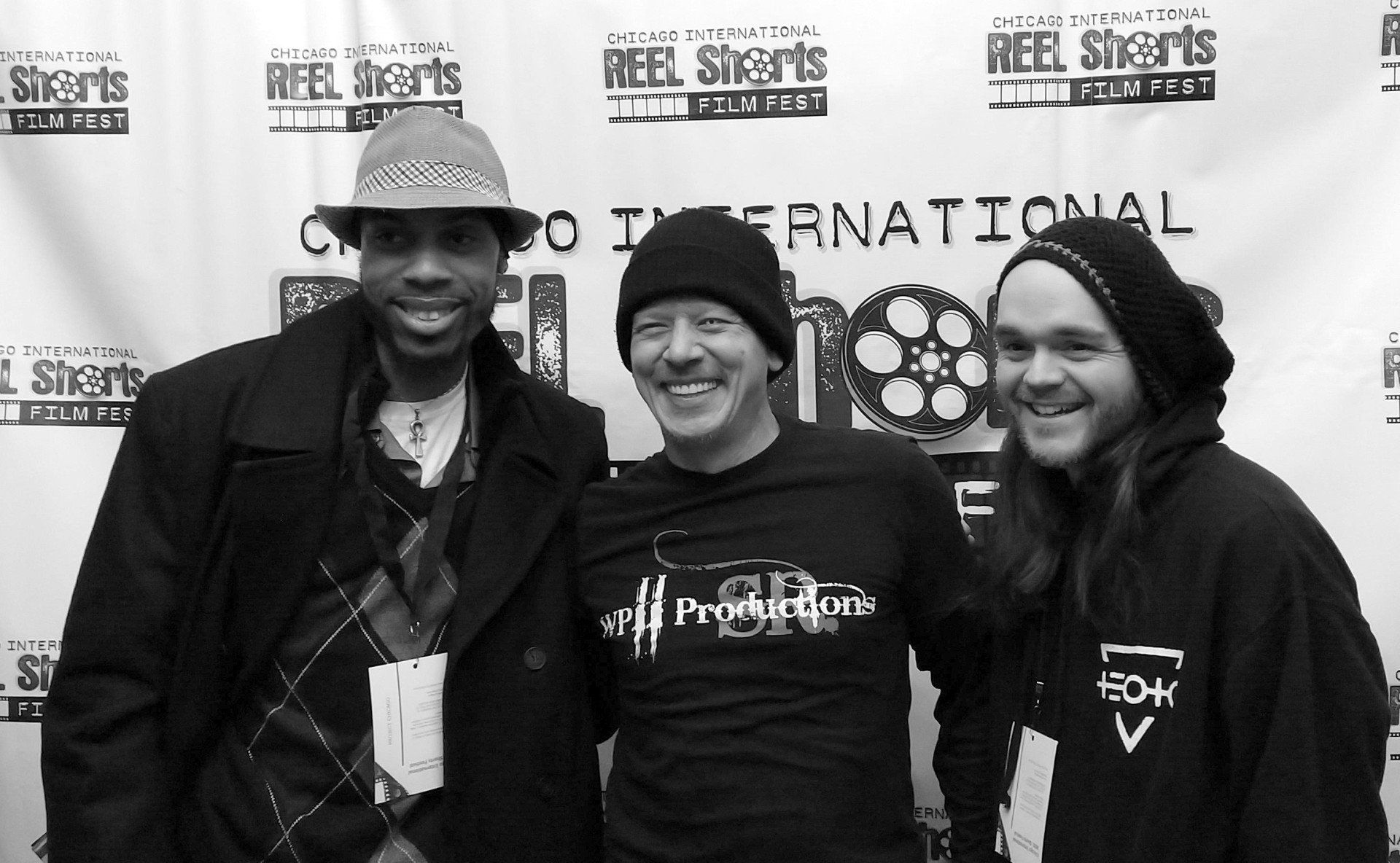 Three men smiling at a film festival. One in a hat and coat, one with a black beanie, and one in a hoodie.