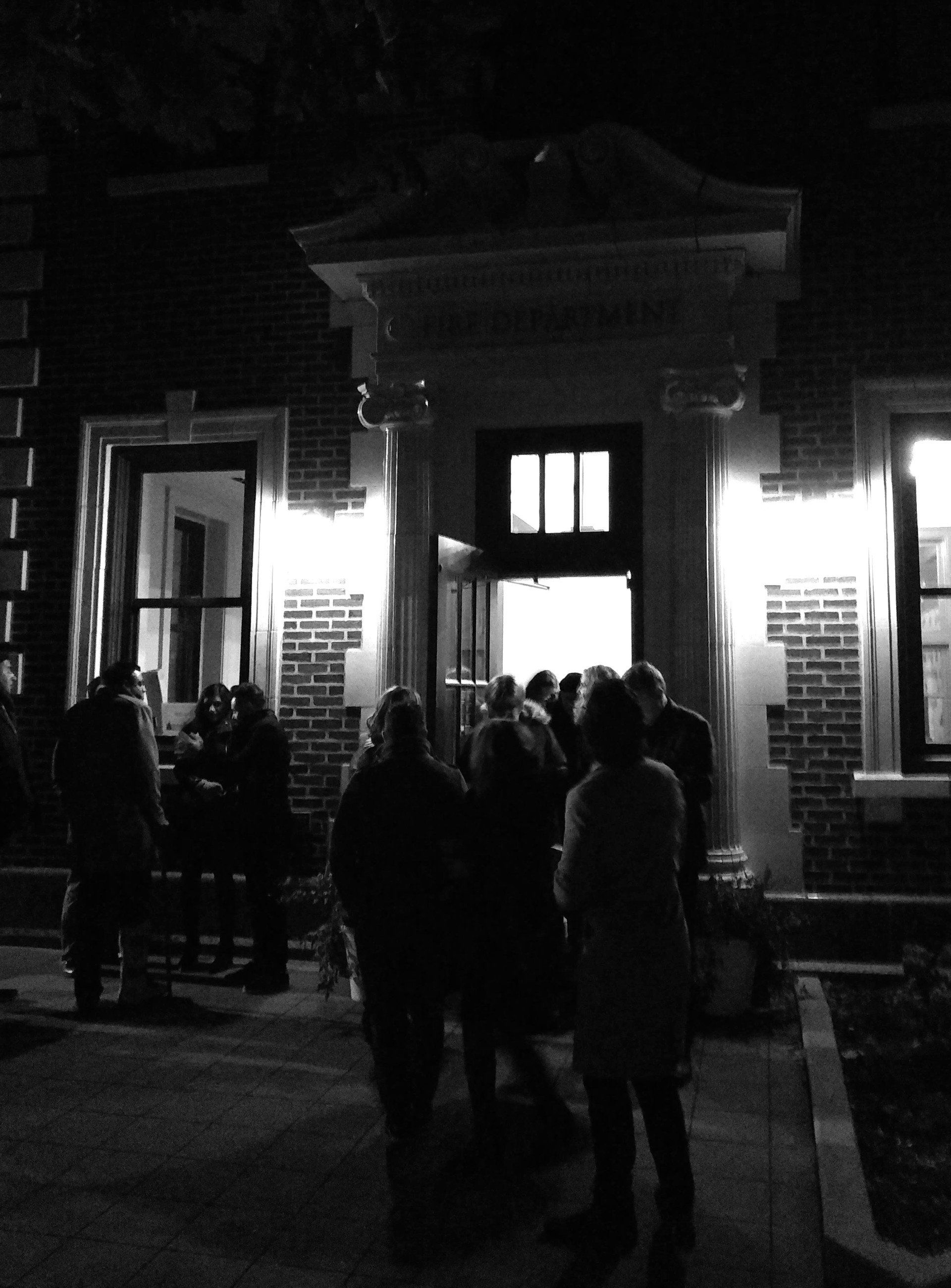 People gather outside a building's entrance, lit by overhead lights. Black and white.