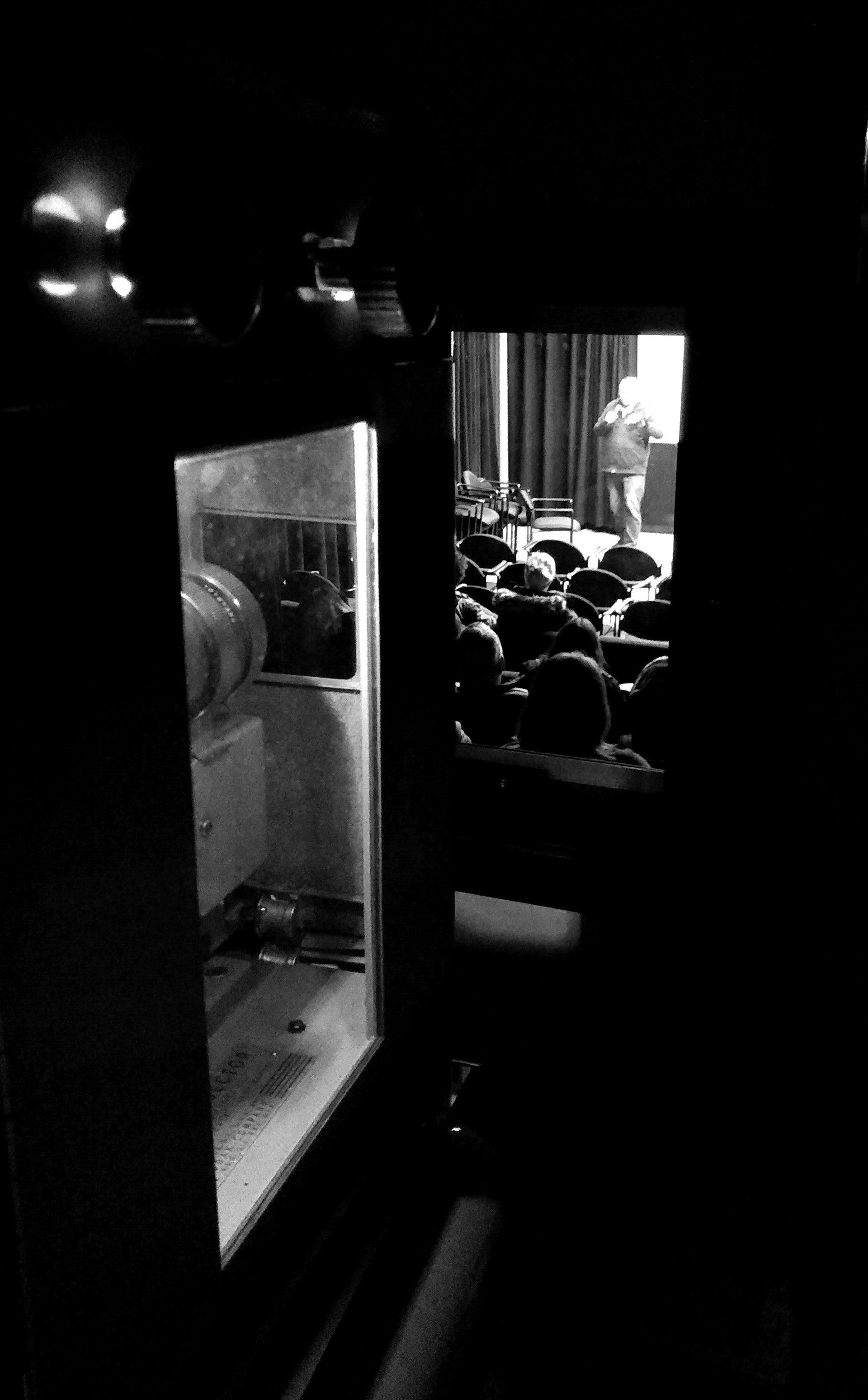 A projection booth window frames a view of a stage and audience. Dark room, black and white.