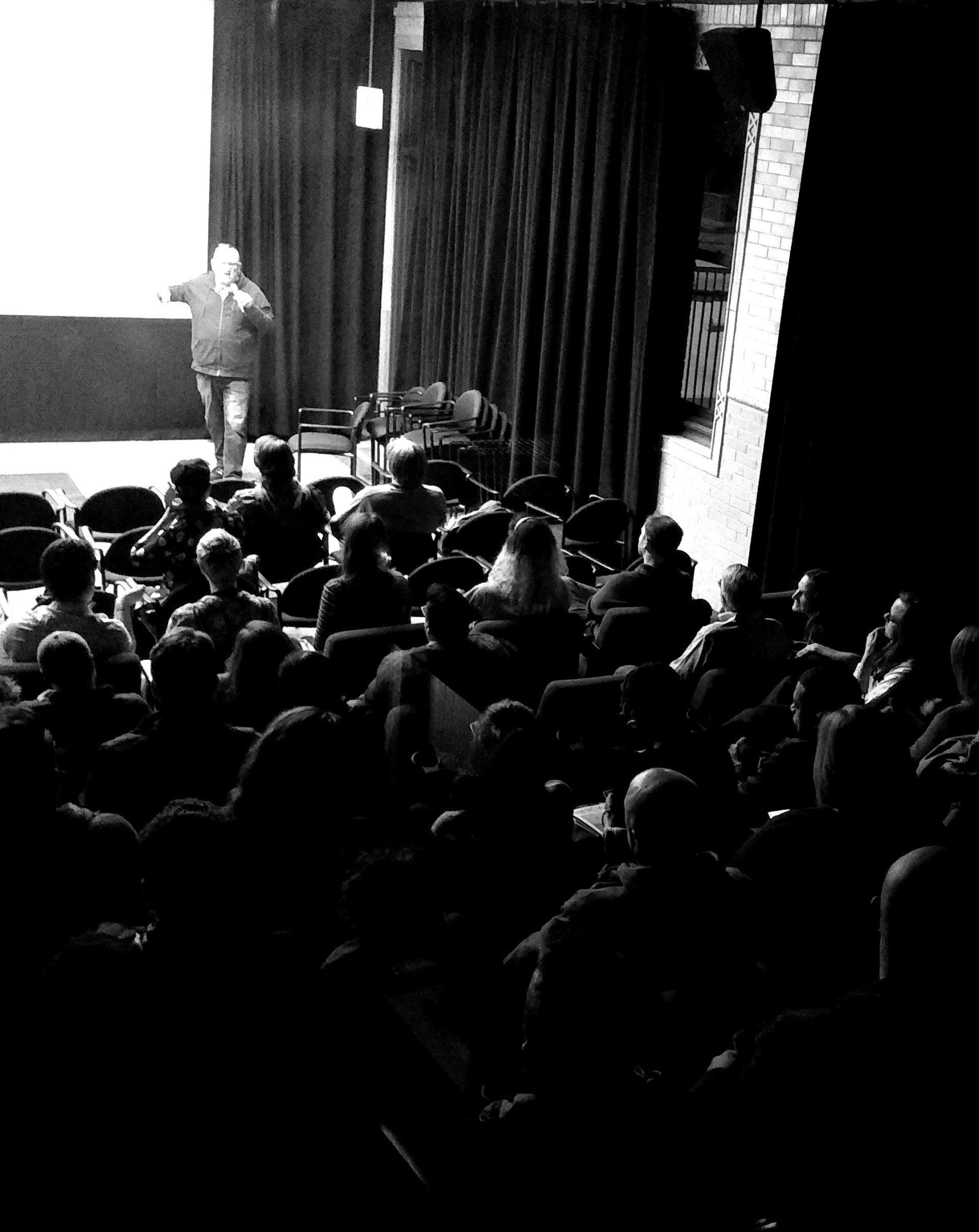 Audience watching a speaker at a dimly lit event in a theater.