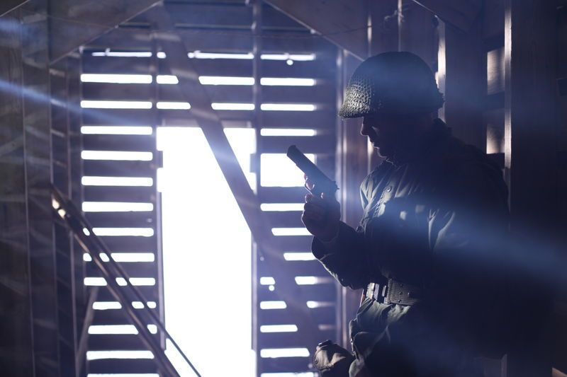 Soldier silhouetted with a pistol inside a wooden structure, bright light through the doorway.