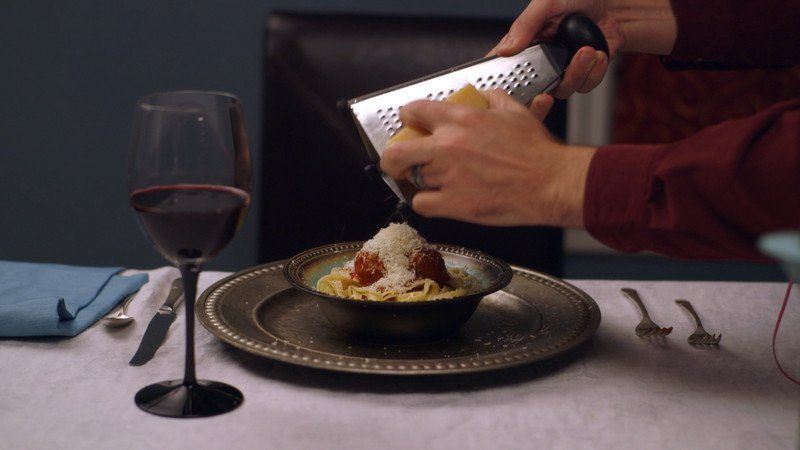 Person grating cheese over pasta with meatballs, alongside wine and silverware.