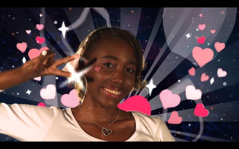 Woman smiles, giving a peace sign with hearts and stars in background, wearing a white shirt and necklace.