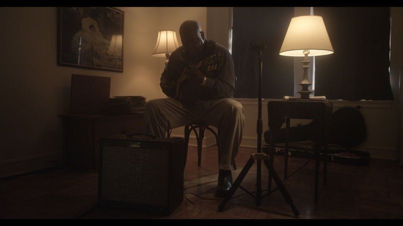 Man sits, holding guitar in dimly lit room, near an amp, a microphone, and lamps.