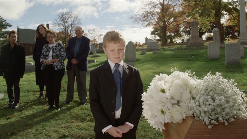 Boy in a suit at a funeral with white flowers; family in background at a graveyard.