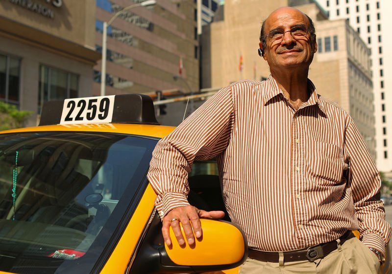 Man in striped shirt leans on a yellow taxi in a city setting, smiling.