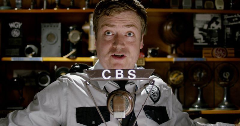 Man in a CBS uniform, eyes wide, standing in front of shelves filled with trophies.