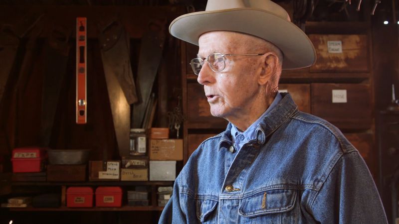 Elderly man in denim shirt and hat speaks inside a workshop with tools and storage shelves.