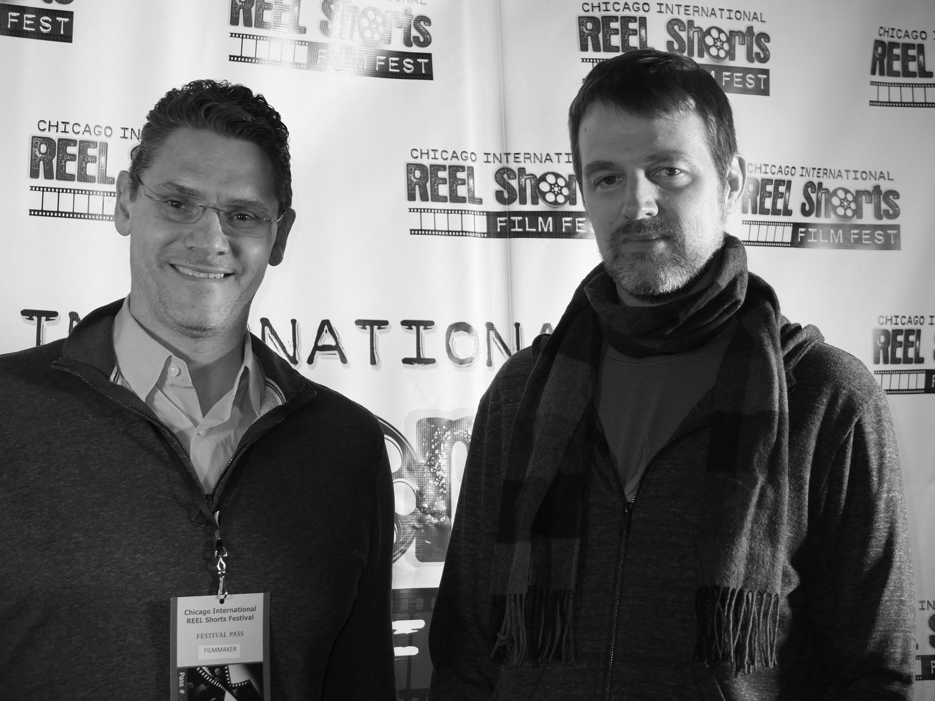Two men pose in front of a Chicago International Reel Shorts Film Fest backdrop. One wears glasses and smiles.