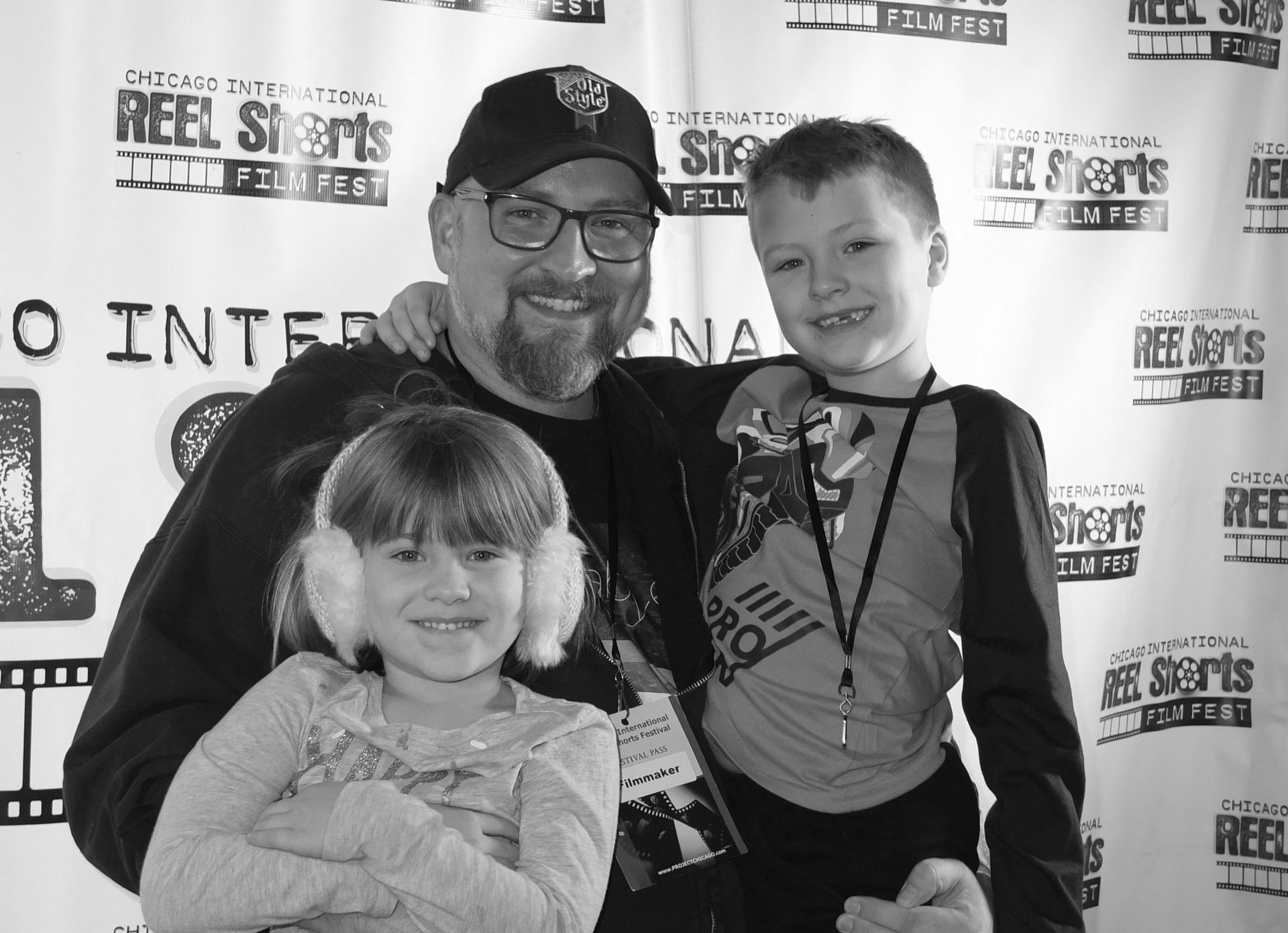 Man with two children smiling at a film festival. He wears a hat and glasses, the girl has earmuffs, and the boy has a lanyard.