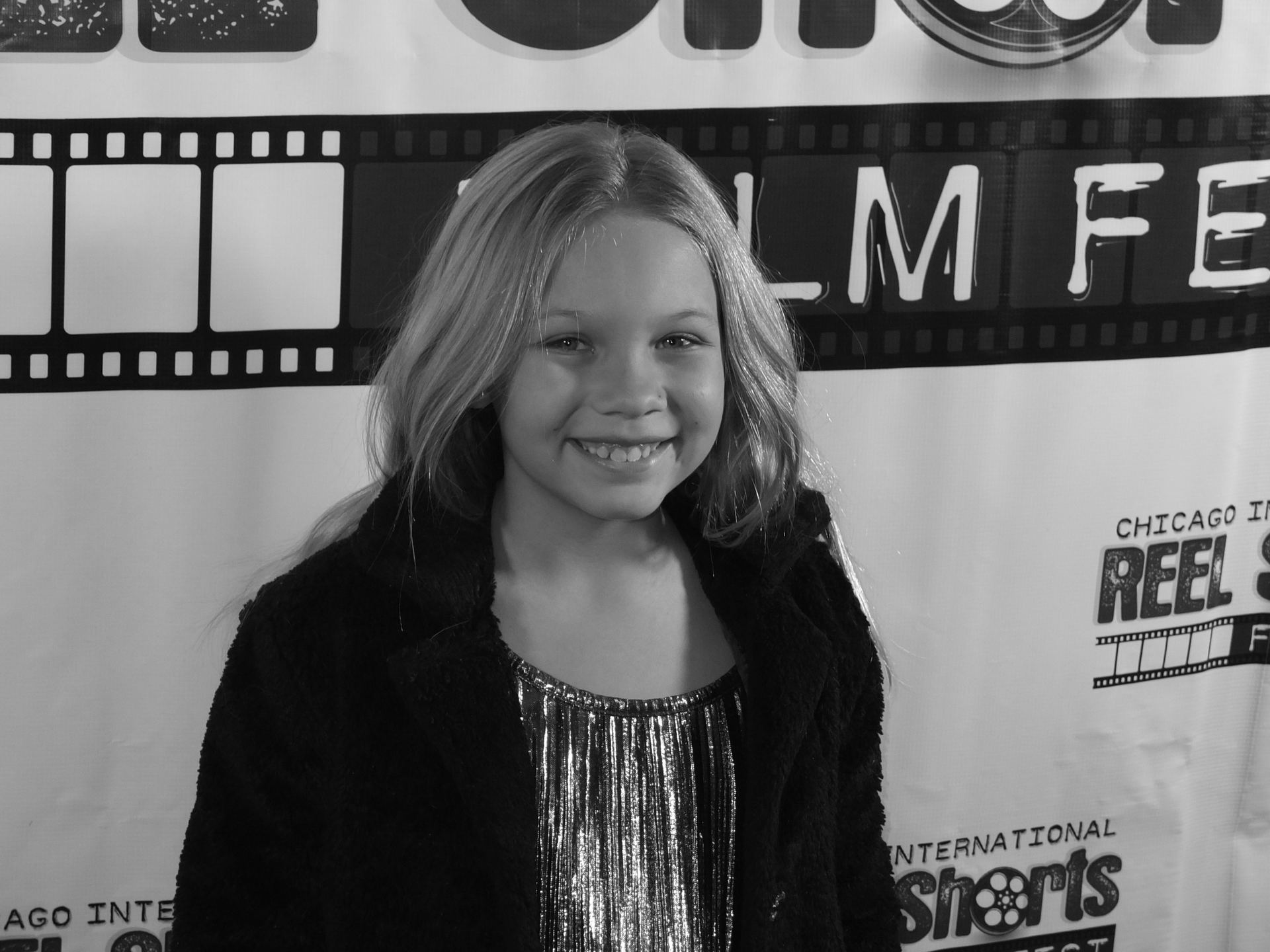Smiling young girl with blonde hair, wearing a sparkly top and coat, at a film festival event.
