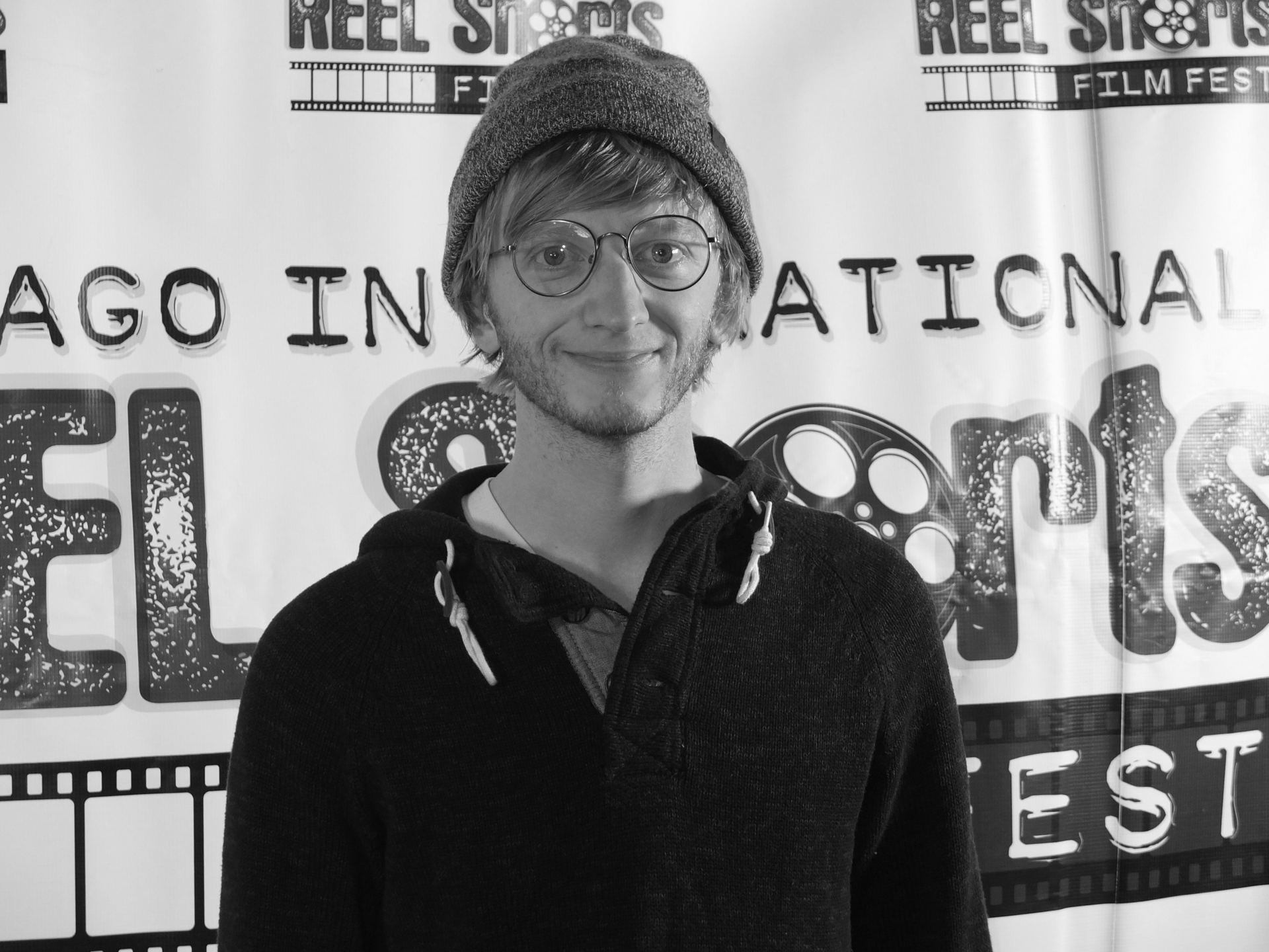 Man with glasses and beanie smiles, posing in front of a festival backdrop.