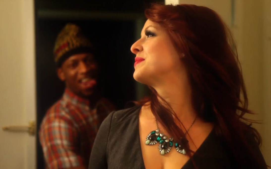 Woman in a dark dress and turquoise necklace, looking up. Man in a hat and plaid shirt, sticking his tongue out in the background.