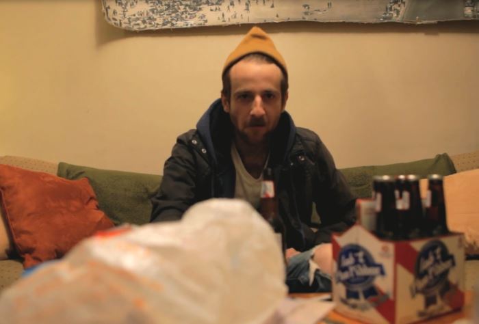 Man with a yellow hat, jacket, and beer sits on a couch with beer bottles. Indoors.
