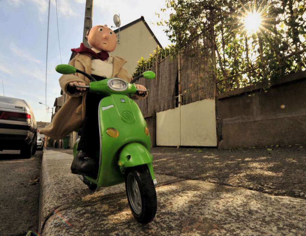 Puppet riding a green scooter down a sidewalk in a sunny residential area.