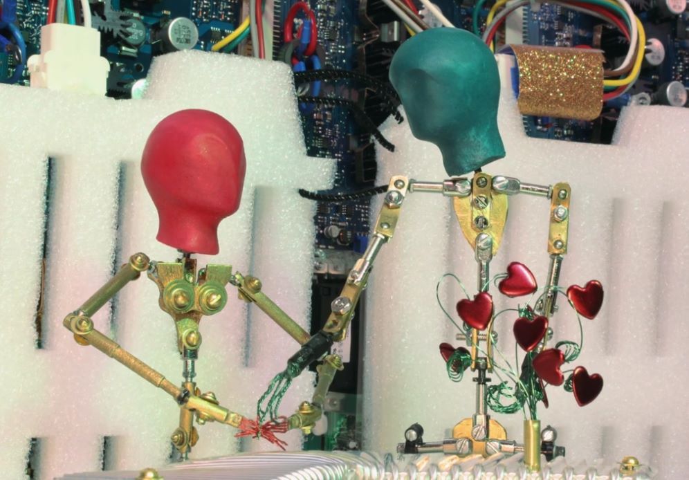 Two robotic figures, red and teal heads, interacting. Teal figure gives a red one red hearts. Background is circuit boards.