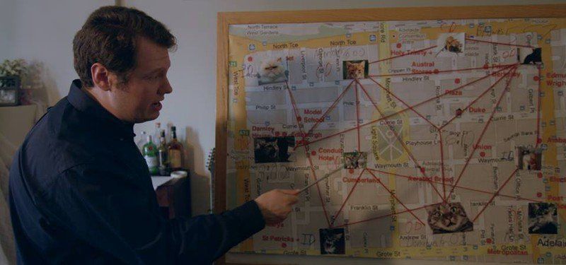 Man pointing at a map with photos and red string, possibly investigating a crime.