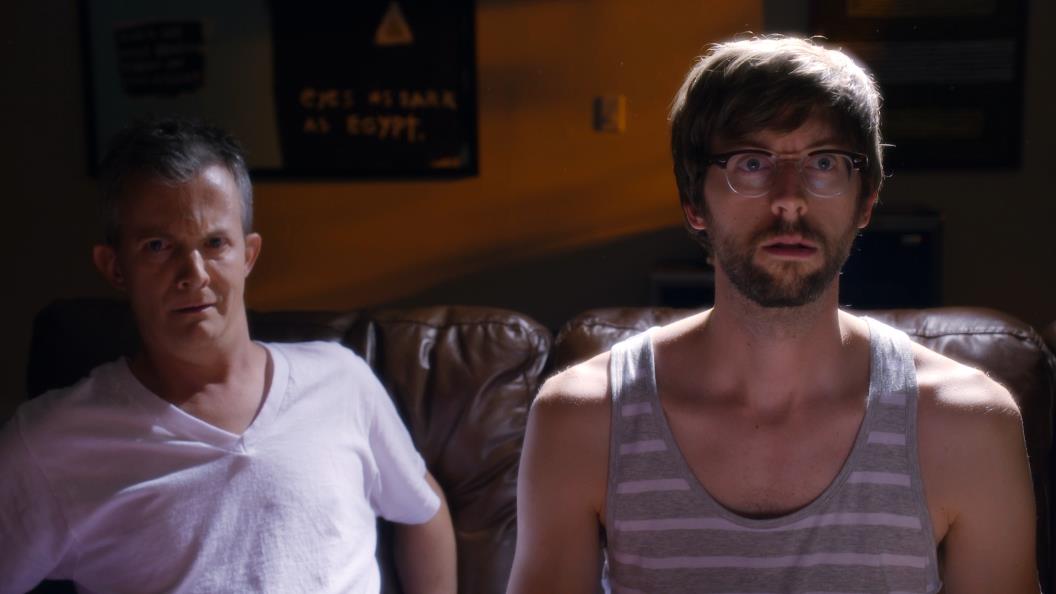 Two men on a couch, one in a white shirt, the other in a tank top, looking surprised. Dim room.