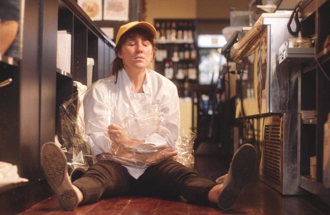 Woman in a restaurant uniform sits on the floor, eyes closed, arms wrapped around something, appearing exhausted.