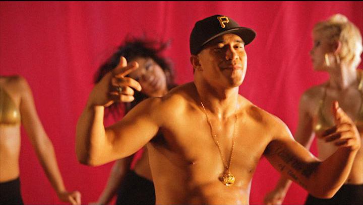 Shirtless man in black cap with gold chain, gestures in front of dancers, red background.