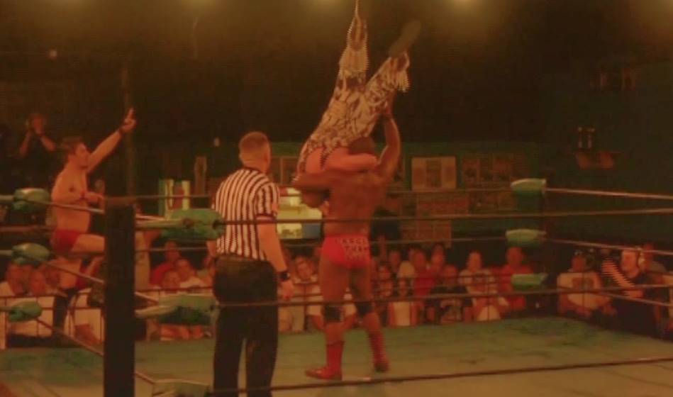 Wrestler lifts another above his head in a wrestling ring. Referee watches. Fans in the background.