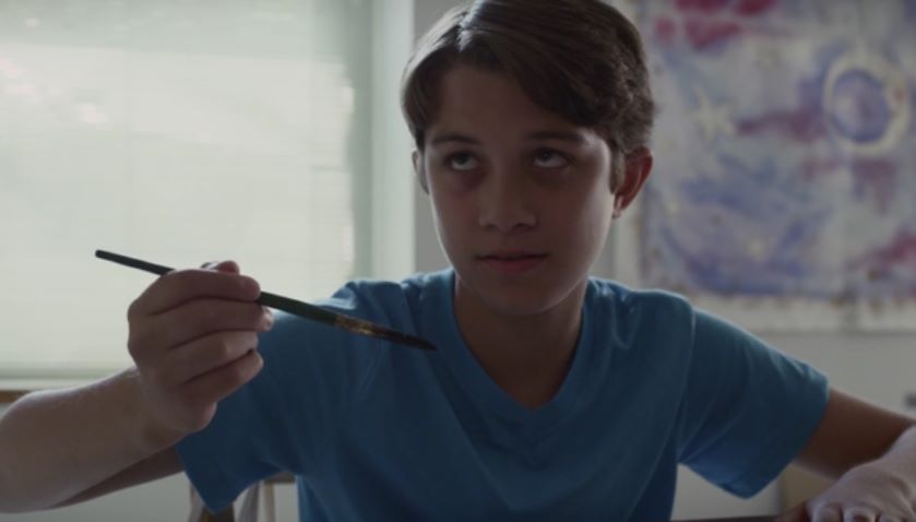 Boy with a paintbrush, gazing intently; painting indoors, blue shirt.