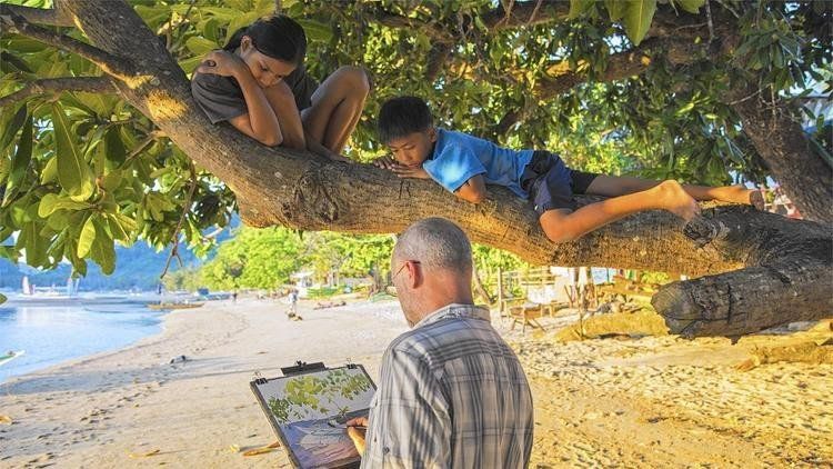 Man paints on beach as two children rest on a tree branch, sunlit by the water.