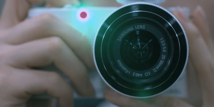 Person holding a white Samsung camera with a lens pointed towards the viewer.