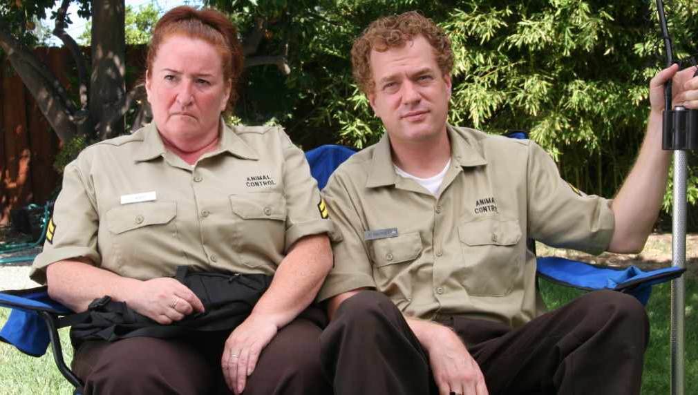 Two people in khaki uniforms sit in blue chairs outdoors; woman frowning, man smiling.