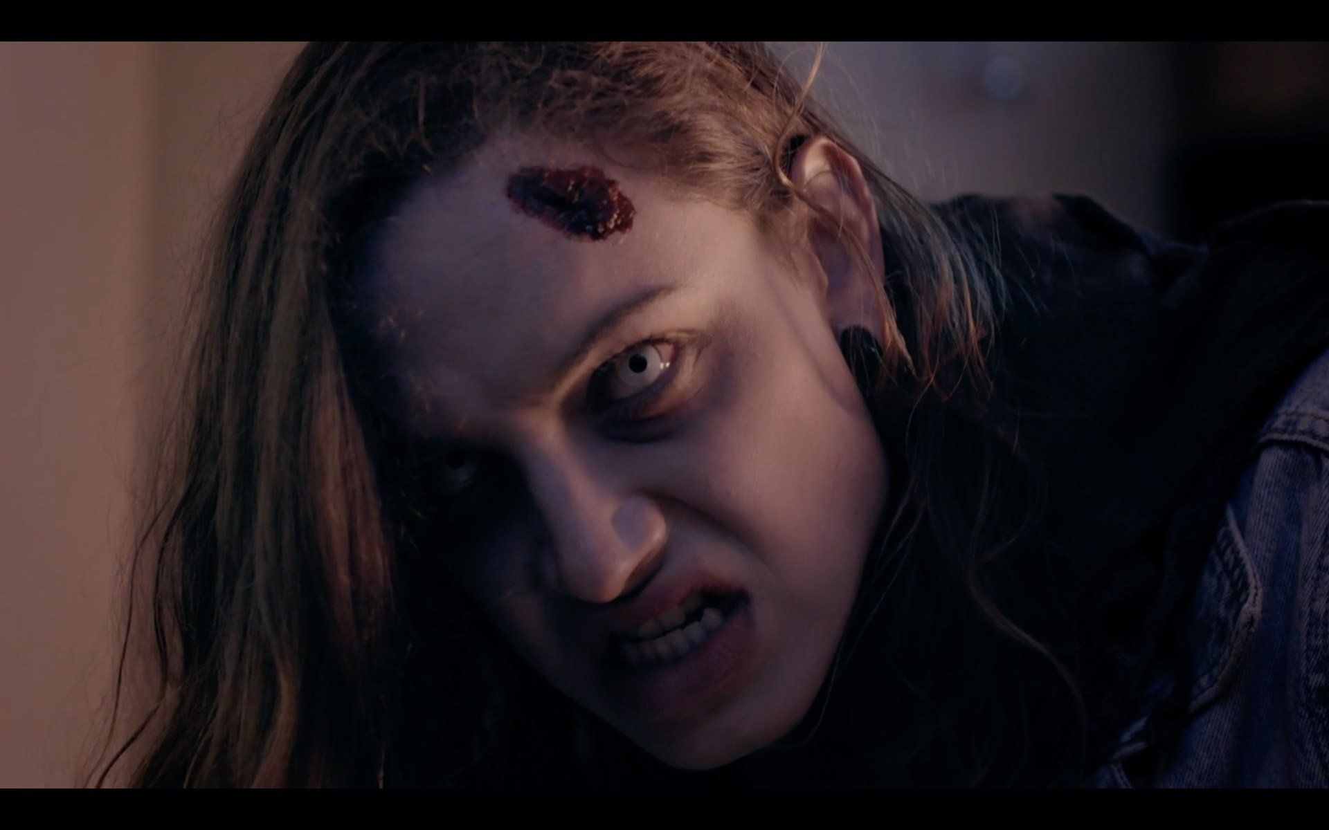 A person with pale skin, white eyes, and a head wound growls in a dimly lit room.