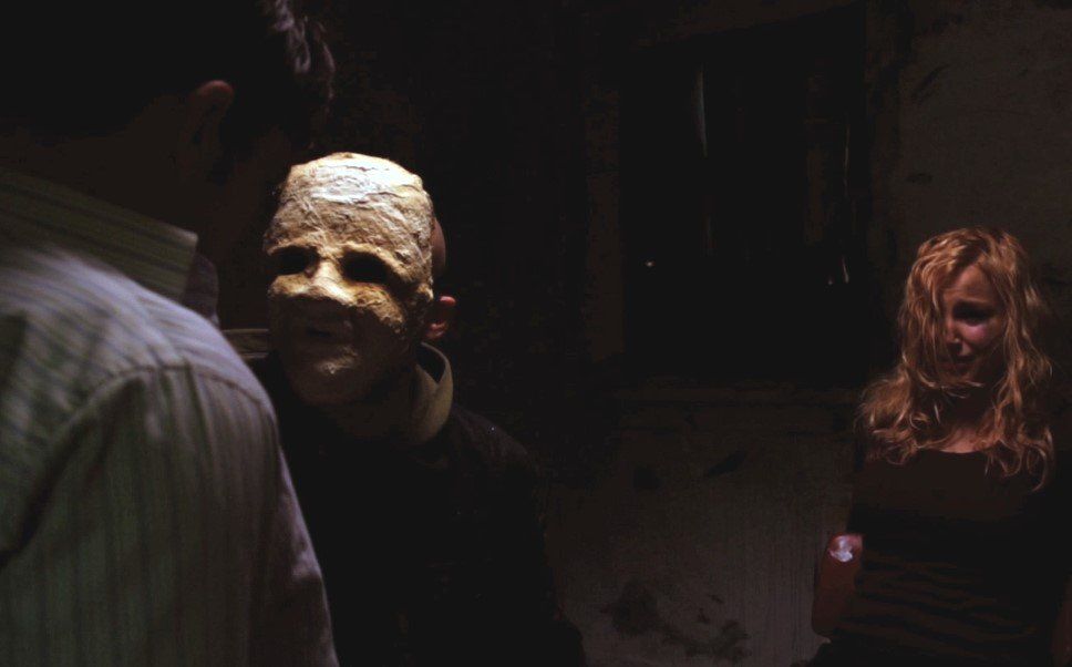 A person in a mask confronts another, as a terrified woman watches in a dark, dingy room.
